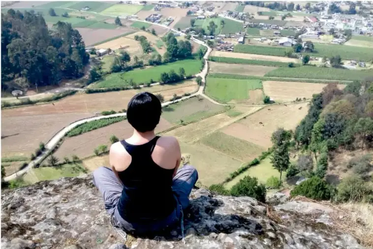 Fotografía de una persona sentada sobre una roca observando un valle con campos agrícolas, caminos, manchones de bosque y un poblado al fondo: ejemplo visual del espacio geográfico como producto de la interacción naturaleza-sociedad.