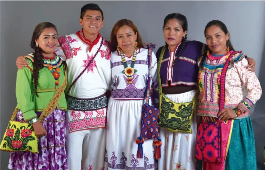 Cinco jóvenes y adultos posando juntos vistiendo coloridas prendas y bordados tradicionales indígenas mexicanos, con bolsas artesanales bordadas.