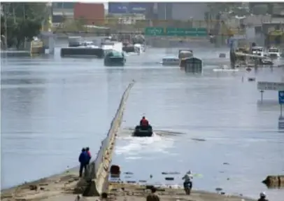 Calle inundada con personas y vehículos; se observa una valla o muro parcialmente sumergido.