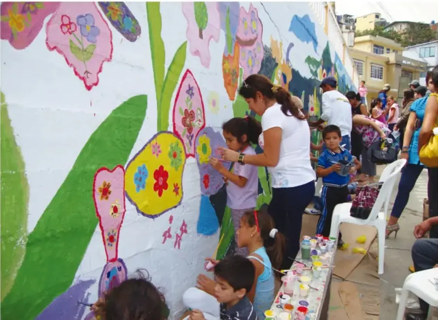 Fotografía de una comunidad pintando un mural colorido con flores y figuras en una pared, con niños y adultos participando.
