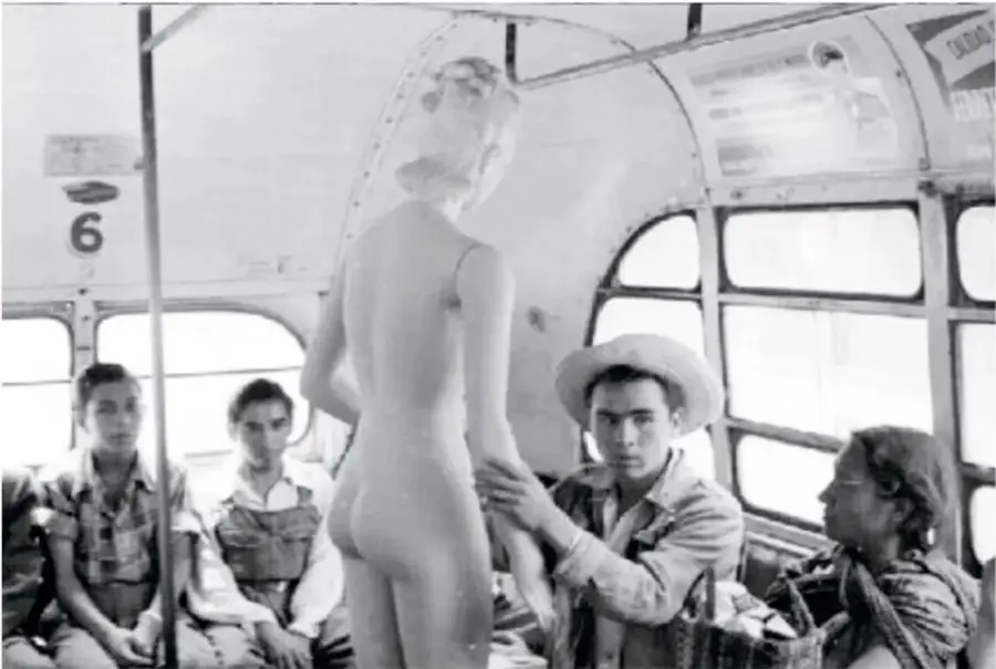 Fotografía en blanco y negro: dentro de un camión urbano, un hombre con sombrero sostiene de la mano a un maniquí femenino desnudo de pie; varios pasajeros observan la escena con curiosidad y asombro.