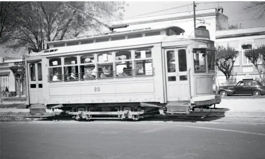 Tranvía antiguo blanco con el número 20, con pasajeros en las ventanas, circulando por una avenida con árboles y un coche antiguo.