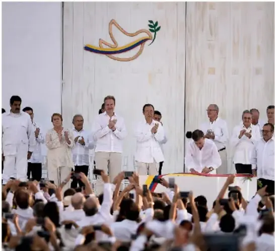 Fotografía de una ceremonia de firma o celebración del Acuerdo de Paz en Colombia, con autoridades y público aplaudiendo frente a una paloma de la paz con los colores colombianos.