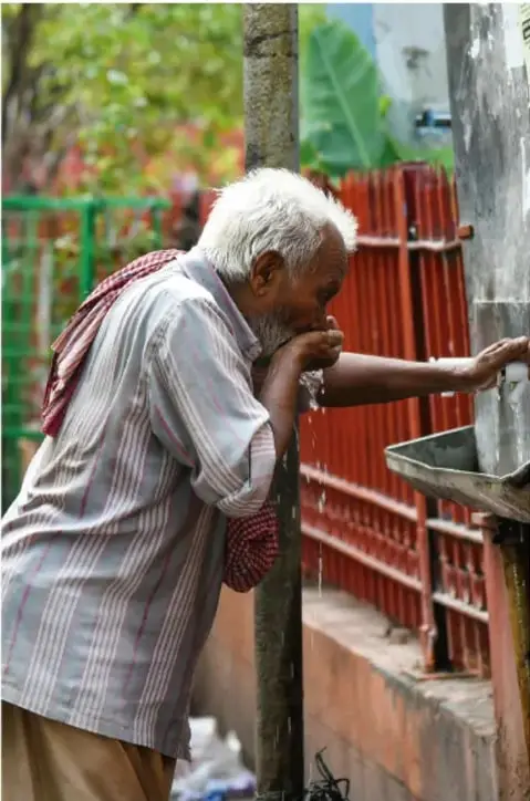 Fotografía de un adulto mayor bebiendo agua directamente de una llave o toma de agua pública en la vía, con cerca de lámina y vegetación de fondo; ilustra el acceso a agua potable como práctica social y tecnológica de la comunidad.