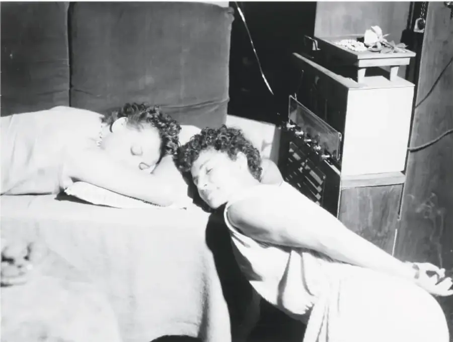 Fotografía en blanco y negro de dos personas dormidas sobre una cama, junto a un aparato eléctrico o radio antiguo.