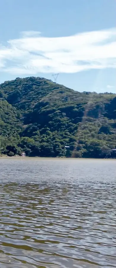 Fotografía del lago de Tequesquitengo con aguas onduladas, cerros verdes y cielo azul con nubes; apenas se ve un pequeño templo a la orilla.