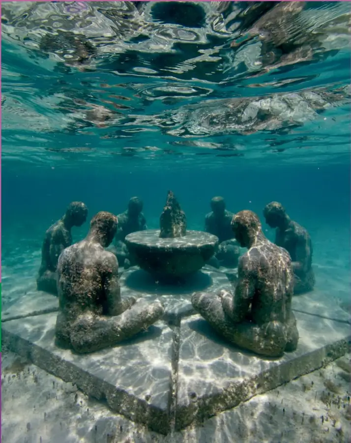 Fotografía subacuática de un conjunto de esculturas de figuras humanas sentadas en círculo alrededor de una vasija o brasero central, colocadas sobre una plataforma cuadrada de piedra en el fondo del mar, con la superficie del agua y la luz solar visibles arriba.