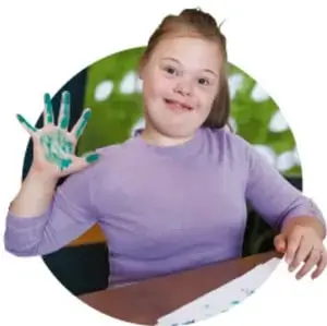Fotografía de una niña sonriendo con las manos pintadas de verde, en un aula con árboles al fondo.
