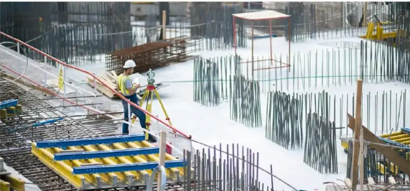 Trabajadores de la construcción en una obra con varillas de acero, andamios y equipo topográfico (teodolito) sobre un trípode.