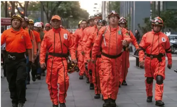 Grupo de personas con trajes naranjas de protección caminando en formación por una calle, probablemente personal de emergencias o brigadas de seguridad.