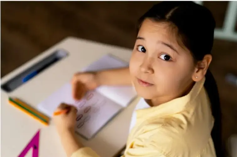 Niña sentada en un escritorio escolar escribiendo en una hoja con lápiz, mirando a la cámara; sobre la mesa hay colores y una regla.