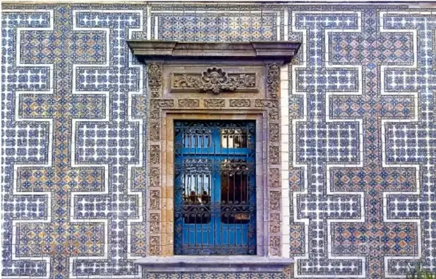 Fotografía de una fachada con azulejos azules y blancos formando un patrón geométrico teselado, con una puerta de hierro forjado en el centro.