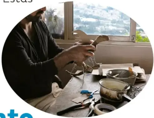 Fotografía en óvalo del mismo artesano pegando tiras de papel de periódico sobre una estructura con alambre, junto a una ventana con vista de la ciudad; hay tijeras, cinta adhesiva y recipientes en la mesa.