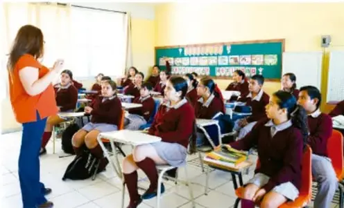 Fotografía de un aula de Telesecundaria donde una docente frente al grupo explica una actividad; el grupo de estudiantes está sentado en sillas observando, con cartelera y materiales educativos al fondo.