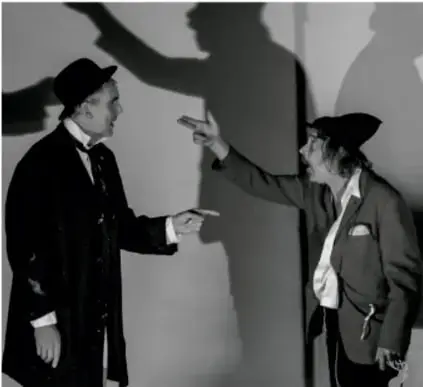 Fotografía en blanco y negro de dos hombres con sombrero señalándose mutuamente frente a una pared con sombras proyectadas, representando la incomprensión en el diálogo.