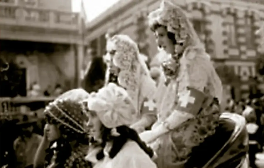 Fotografía en sepia de mujeres voluntarias de la Cruz Blanca a bordo de un vehículo, ca. 1915; llevan mantilla, delantal y brazaletes con cruz roja/blanca; el fondo muestra una calle con edificios y espectadores. © INAH, SINAFO, FN.MX.
