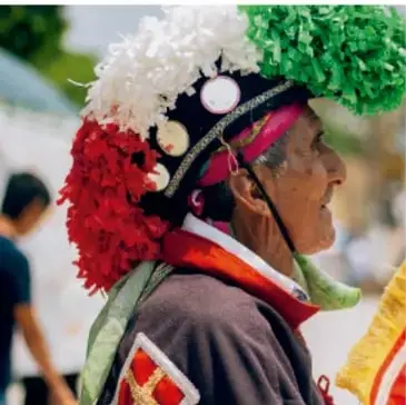 Persona en traje tradicional con penacho de flores blancas, verdes y rojas, en un contexto de fiesta comunitaria.