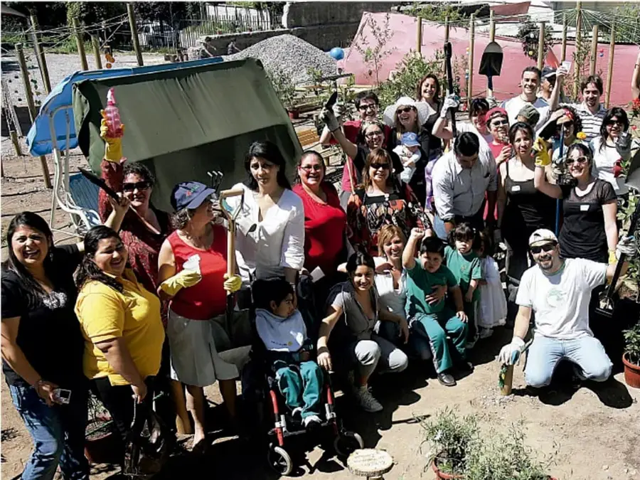 Fotografía de un grupo numeroso de miembros de la comunidad (adultos, niños y una persona en silla de ruedas) sonrientes, con guantes y herramientas de jardinería, en un espacio al aire libre con plantas y estructuras sencillas; representa un proyecto comunitario ejecutado con participación colectiva.