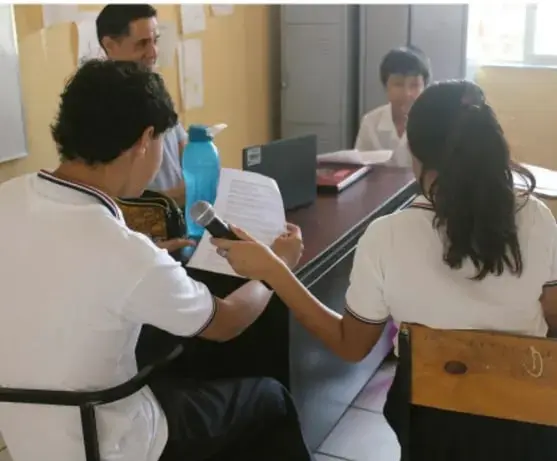 Fotografía de estudiantes en un aula trabajando con equipo de cómputo y un micrófono, preparando la presentación de su poemario.