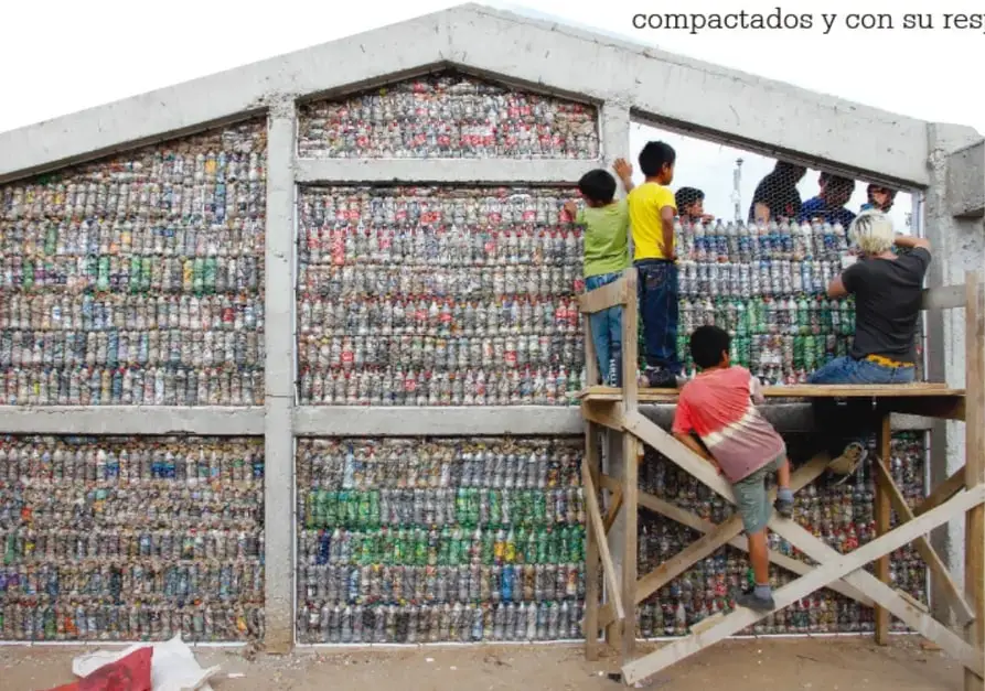 Fotografía de un muro construido con cientos de botellas plásticas rellenas (ecoladrillos), con varios niños y adultos trabajando sobre andamios de madera.