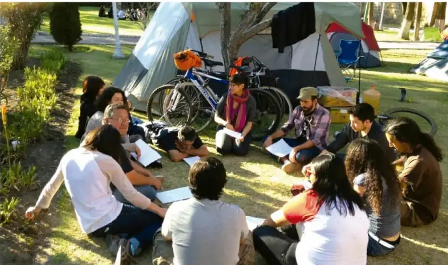 Fotografía de un grupo de jóvenes sentados en círculo sobre el pasto, junto a carpas y bicicletas, tomando notas en cuadernos; representan la planeación y comunicación en equipo.