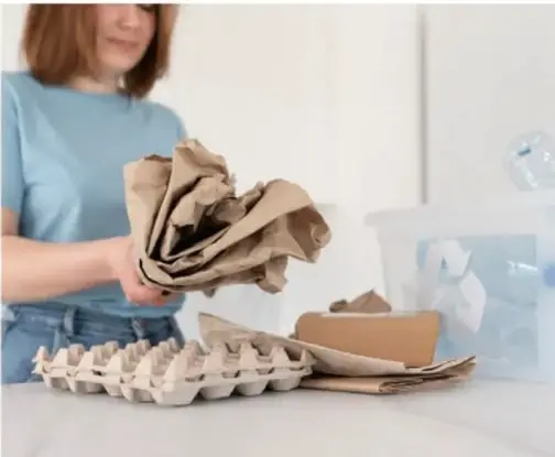 Mujer que clasifica residuos reciclables (papel, cartón, bandejas de huevo) en contenedores sobre una mesa.