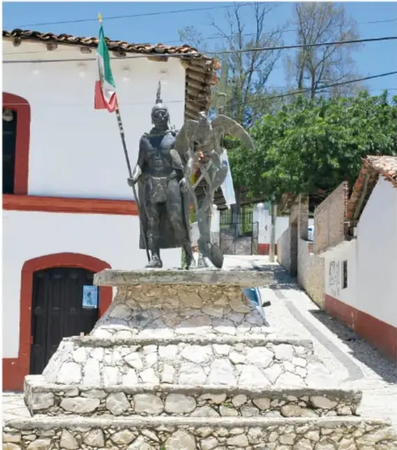 Estatua de Cuauhtémoc con bandera mexicana en una calle del pueblo de Ixcateopan, con muros blancos y tejas rojas.