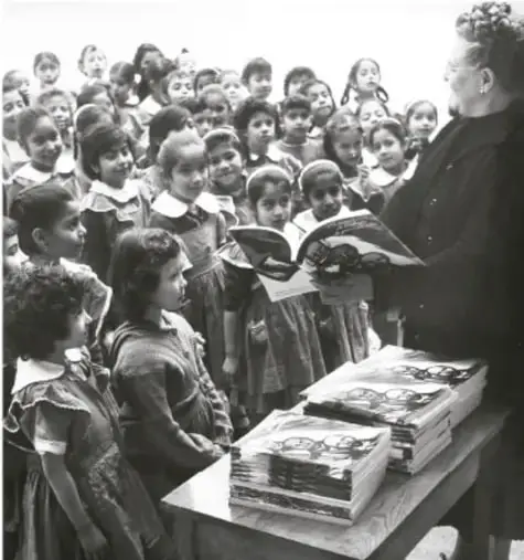 Fotografía en blanco y negro de una maestra mostrando un libro a un grupo de niñas y niños en un salón de clases.