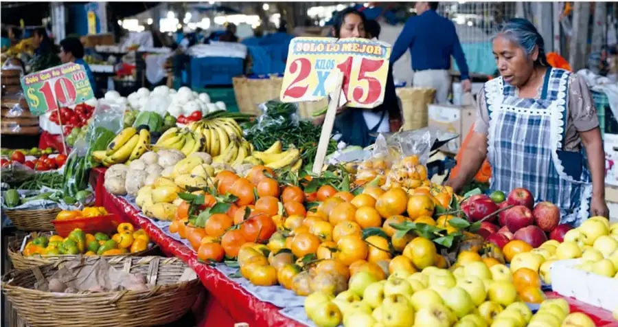 Puesto de mercado con frutas y verduras, carteles de precios en pesos y una vendedora.