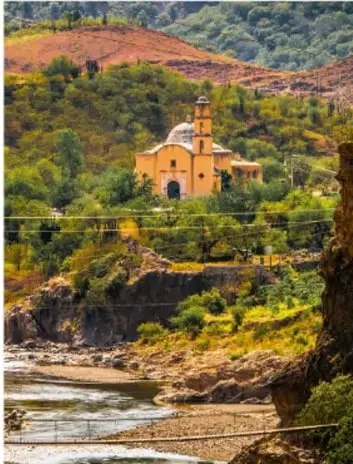 Fotografía de una iglesia de misión con fachada ocre y torre campanario, situada en un cañón verde con río; ambiente serrano del norte de México.