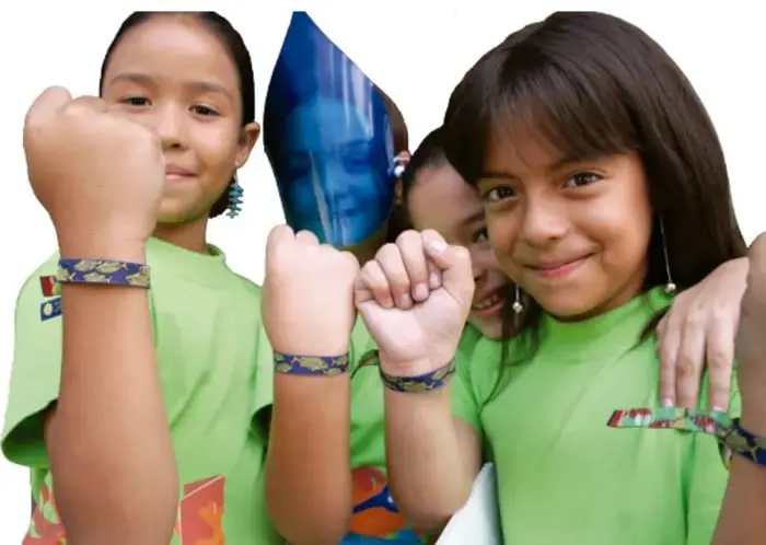 Tres niñas con playeras verdes levantan el puño y sonríen; una sostiene un globo azul. Representan la unión y la participación comunitaria.