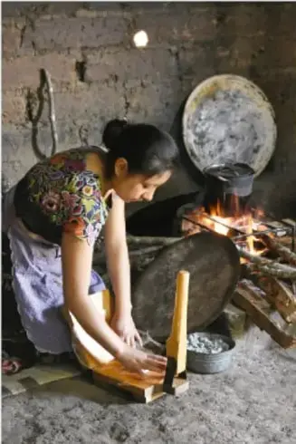 Fotografía de una mujer indígena en un interior de paredes de adobe, arrodillada junto a un fogón tradicional con fuego encendido entre piedras, avivando la llama con un abanico de madera; a su lado hay utensilios de cocina y un comal.