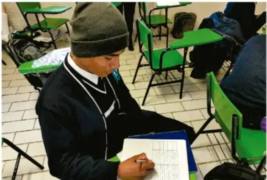 Fotografía de un estudiante de Telesecundaria con gorro y uniforme, sentado en su silla-escribanía verde, escribiendo en su cuaderno; otros pupitres verdes y compañeros se ven alrededor. Crédito: Luis Eduardo García Nieto, INEE.