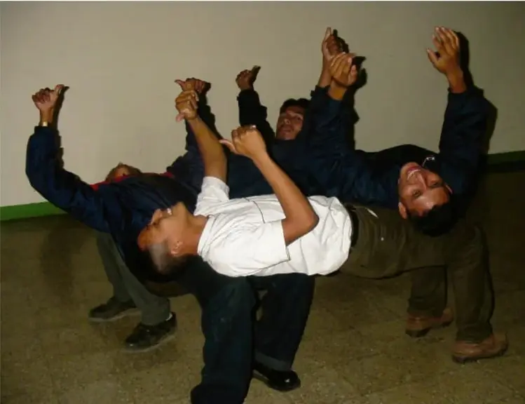Fotografía de un grupo de estudiantes realizando una dinámica de confianza: varios alumnos sostienen horizontalmente a sus compañeros con los brazos extendidos, en un espacio cubierto.