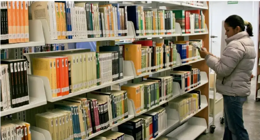 Fotografía de una estudiante consultando libros en los estantes de una biblioteca escolar.