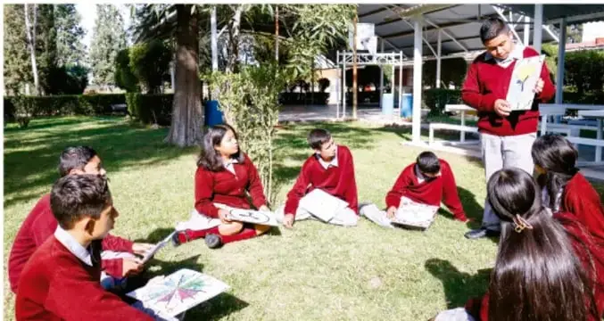 Fotografía de un grupo de estudiantes de secundaria con uniforme rojo sentados en círculo sobre el pasto, leyendo y comentando sus trabajos, mientras un compañero de pie lee en voz alta.