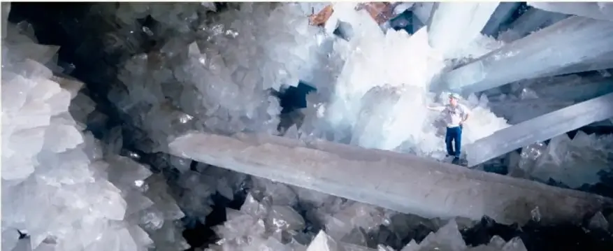 Fotografía de gran formato del interior de la Cueva de los Cristales de Naica: enormes vigas de cristales de yeso translúcidos, con una persona de pie sobre uno de ellos para mostrar la escala.