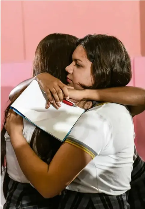 Fotografía de dos estudiantes abrazándose, una de ellas sostiene un cuaderno; fondo rosa, posiblemente ilustrando apoyo entre compañeras frente al acoso.