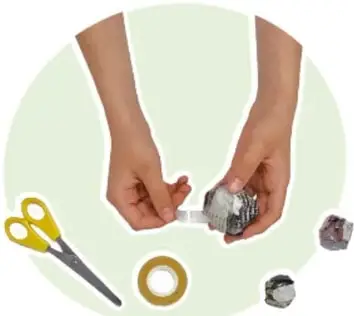 Fotografía de manos sosteniendo un tubo de cartón de papel higiénico sobre un pedazo de cartón café; alrededor hay tijeras, un plumón negro, cinta y bolas de papel.