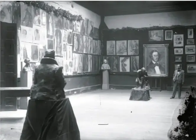 Fotografía en blanco y negro de una sala de museo con paredes cubiertas de cuadros y esculturas; en primer plano, una figura de espaldas con abrigo observa la sala.