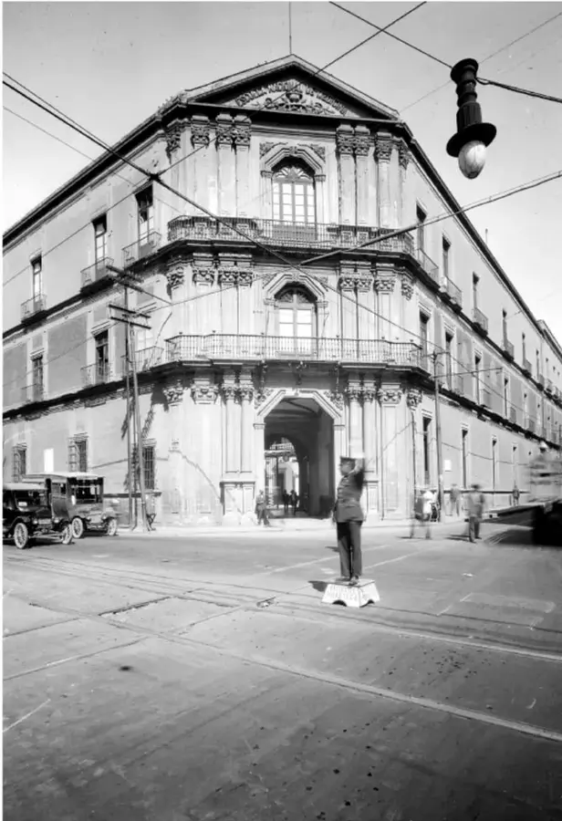 Fotografía en blanco y negro de un edificio antiguo de esquina con balcones y arcos, cables de luz aéreos, un poste con lámpara colgante, un automóvil de época estacionado y una persona de uniforme con los brazos extendidos dirigiendo el tránsito en una calle con vías de tranvía.