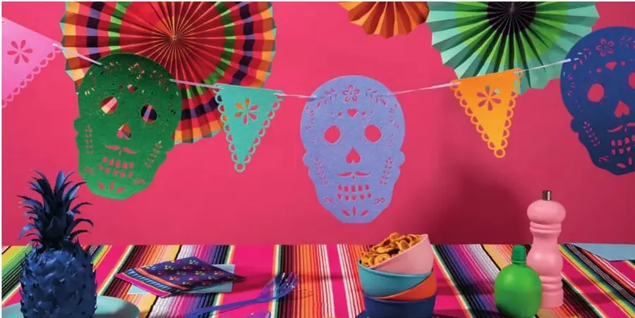 Fotografía decorativa con calaveras de papel picado (verde, púrpura, azul), abanicos de papel coloridos, una piña azul y una mesa con tazones, en alusión al Día de Muertos mexicano.