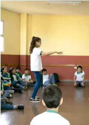 Fotografía de un taller de teatro: una niña de pie frente al grupo lee o actúa, mientras sus compañeros sentados en el suelo la observan.