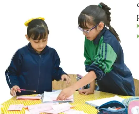 Dos niñas de primaria trabajan en una mesa con hojas y colores; una de ellas señala un papel mientras la otra escribe.