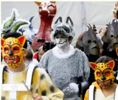 Fotografía de danzantes con máscaras tradicionales de jaguar y otros animales y un personaje con vestimenta gris y pintura facial, en una celebración comunitaria mexicana.