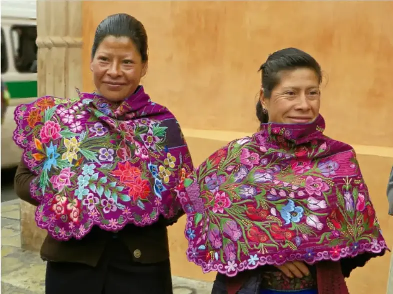 Fotografía de dos mujeres indígenas mexicanas con rebozos bordados de colores vibrantes (flores multicolores sobre fondo morado), mostrando manifestaciones artísticas y culturales tradicionales.