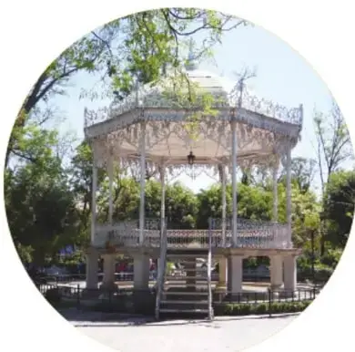 Fotografía del kiosco blanco del Jardín de San Marcos, de dos niveles con escalinatas, rodeado de árboles y bajo el sol.