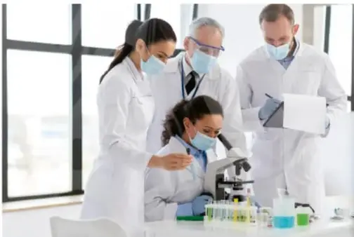 Fotografía de un equipo de cuatro científicos con batas blancas y mascarillas en un laboratorio; uno observa al microscopio y otros revisan muestras y anotan datos.
