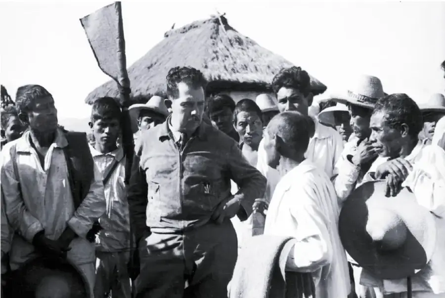 Fotografía en blanco y negro de Lázaro Cárdenas rodeado de campesinos, uno de ellos sostiene un sombrero, en un entorno rural con una choza de techo de paja al fondo.