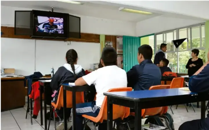 Fotografía de un aula donde estudiantes vestidos con uniforme observan una presentación en una pantalla de TV montada en la pared, mientras otros expositores de pie frente a la comunidad en el fondo derecho.